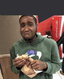 a man wearing a green hoodie is eating a sandwich in front of a coca cola machine