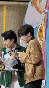 a man in a brown jacket stands next to another man in a green jacket holding a microphone ..