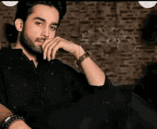 a man with a beard is sitting on a couch with his hand on his chin and wearing a watch .