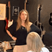 a woman in a black shirt stands in front of a wall of guitars
