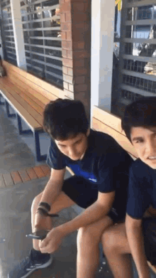 two boys are sitting on a wooden bench looking at their phones