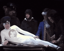 a man in a white suit is laying on a table surrounded by people