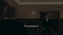 a man in a suit is talking to another man in a dark room and the man in the suit says you know it