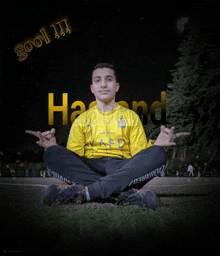 a young man in a yellow shirt sits in a lotus position in front of a sign that says hool