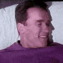 a man in a purple shirt is laying in a bed with his eyes closed and smiling .