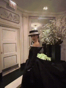 a woman in a black dress and hat stands in a hallway with a red exit sign above her head