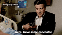 a man in a suit is sitting in a hospital bed looking at his phone and says `` here have some concealer '' .