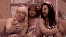 three men dressed as women are sitting on a couch taking a selfie with their cell phones .