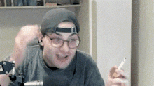 a man wearing a hat and glasses is holding a cigarette