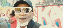 a man wearing sunglasses and a hat is standing in front of a wall with graffiti .