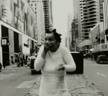 a black and white photo of a woman standing on a city street covering her mouth with her hand .