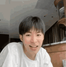 a young man in a white t-shirt is smiling and looking at the camera in a living room .