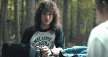 a man with long curly hair wears a hellfire club shirt