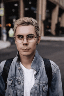 a young man wearing glasses and a denim jacket stands in front of a building