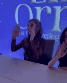 two women are sitting at a table with their hands in the air in front of a sign that says orion .