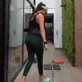 a woman in a black tank top and green leggings is standing next to a barbell in a hallway .