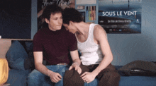 two men are sitting on a bed in front of a movie poster that says sous le vent