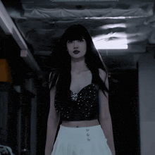 a woman in a black crop top and white skirt stands in a dark room