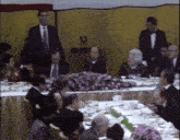 a man in a tuxedo stands in front of a table full of people