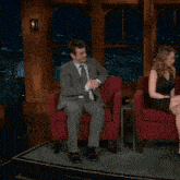 a man in a suit and tie sits next to a woman in a black dress in red chairs
