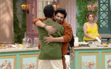 a man in a green shirt is hugging another man in an orange shirt in a kitchen