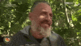 a man with a beard and a givova jacket is standing in the woods
