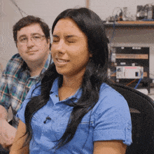 a woman in a blue shirt sits in a chair with her eyes closed and a man in a plaid shirt sits behind her