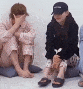two women are sitting on the floor covering their faces with their hands . one of the women is wearing a hat .