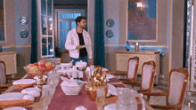 a man in a white jacket is standing in front of a dining room table with plates and bowls on it .