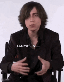 a young man in a suit and black shirt is sitting in a chair with his hands on his chest .
