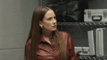 a woman in a red leather shirt is standing in front of a metal shelf with cases on it .