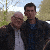 two men standing next to each other with one wearing glasses and the other wearing a jacket