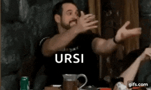 a man with a beard is sitting at a table making a funny face and pointing at the camera .
