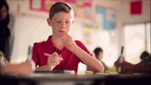 a boy in a red shirt is sitting at a desk in a classroom eating a candy bar .