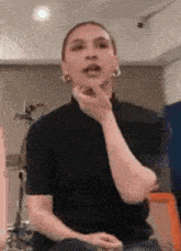 a woman in a black shirt and hoop earrings is sitting in front of a microphone with her hand on her chin .
