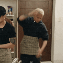 a woman in a plaid skirt is dancing in a kitchen with a man in a black shirt standing behind her .