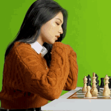 a woman in an orange sweater sits at a table with a chess board in front of her