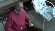 a man in a pink sweater is sitting on a porch with a chair in the background .