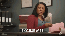 a woman is sitting at a desk holding a piece of paper and saying `` excuse me ? ''