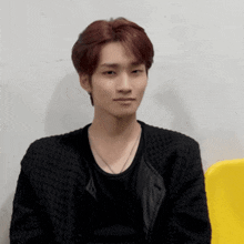 a young man with red hair is wearing a black jacket and sitting on a yellow chair