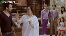 a woman in a white saree is standing in front of a group of people in a living room .