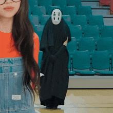a girl in overalls stands next to a person wearing a black mask