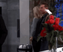 a man and a woman are standing next to a vase of red roses in a kitchen .