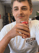 a young man wearing a white volcom shirt is holding a can of soda