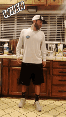 a man in a white shirt and black shorts stands in a kitchen with the word when written above him