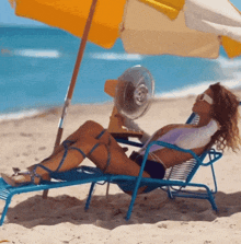 a woman sits on a beach chair under an umbrella while holding a fan