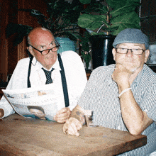 two men are sitting at a table reading a newspaper which says ' clockham pictular ' on the front