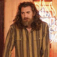 a man with long hair and a beard is standing in front of a neon sign