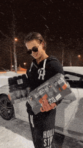 a man wearing sunglasses and a hoodie with the letter h on it is holding a stack of bottles of nocco