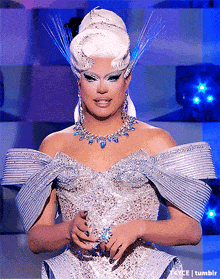 a drag queen wearing a white dress and a necklace is standing in front of a tumblr logo